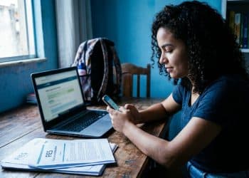 Estudante acessa o Desenrola Fies pelo celular para renegociar a dívida do Fies, com documentos do financiamento sobre a mesa.