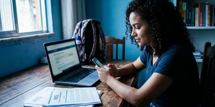 Estudante acessa o Desenrola Fies pelo celular para renegociar a dívida do Fies, com documentos do financiamento sobre a mesa.