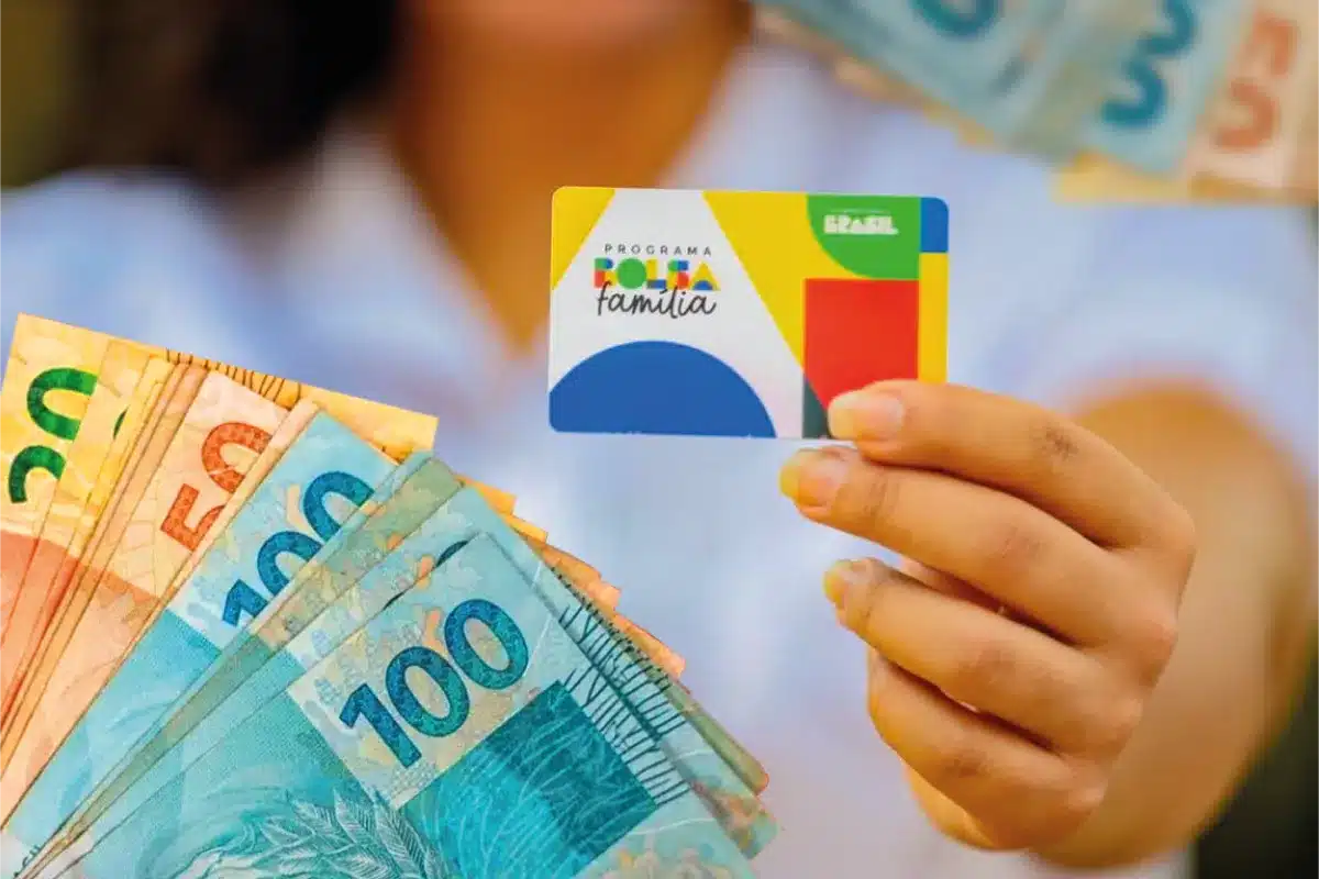 Mão segurando o cartão do Programa Bolsa Família ao lado de um leque de cédulas de R$ 50 e R$ 100, em referência aos valores adicionais pagos no benefício de junho de 2026