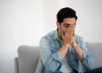 Homem em ambiente doméstico demonstrando sinais de ansiedade e tensão emocional ao cobrir o rosto com as mãos.
