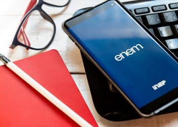 Celular exibindo a logo do Enem na tela ao lado de teclado, óculos, caderno e lápis, em referência ao resultado do atendimento especializado do Enem 2026