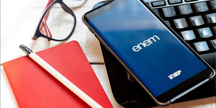 Celular exibindo a logo do Enem na tela ao lado de teclado, óculos, caderno e lápis, em referência ao resultado do atendimento especializado do Enem 2026