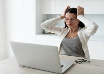 Mulher com as mãos na cabeça diante do notebook representa o estresse e o esgotamento causados pela autocobrança