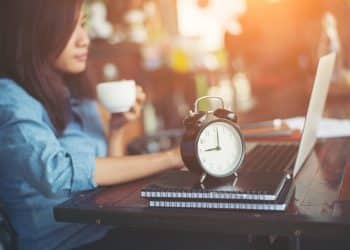 Mulher toma café ao lado de relógio despertador e notebook sobre mesa de trabalho