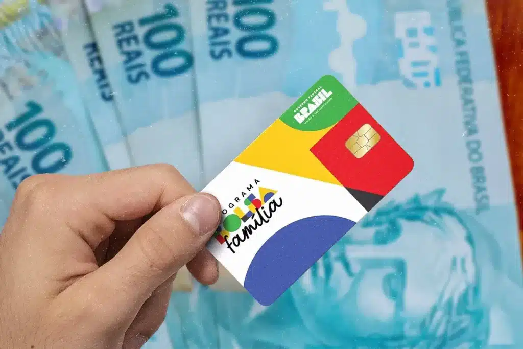 Cartão do Bolsa Família em destaque sobre notas de R$ 100, representando os adicionais pagos às famílias beneficiárias.