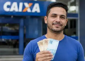 Homem brasileiro em frente à agência da Caixa Econômica Federal segurando dinheiro, representando liberação de saque de até R$ 6.220.