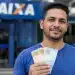 Homem brasileiro em frente à agência da Caixa Econômica Federal segurando dinheiro, representando liberação de saque de até R$ 6.220.