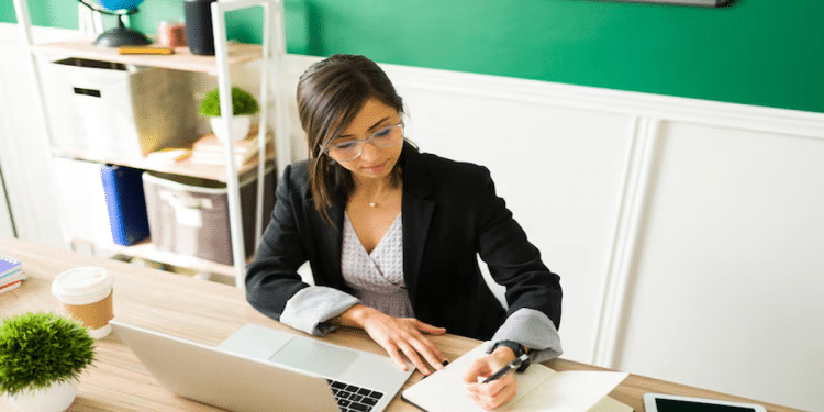 Mulher de óculos e blazer escreve em caderno ao lado do notebook