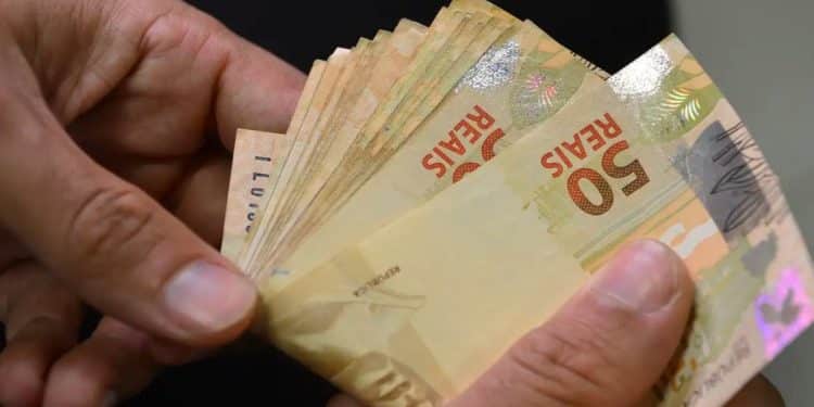 Mãos contando cédulas de R$ 50, representando o pagamento de R$ 2.216 que une o abono salarial e o Cashback do Imposto de Renda em 15 de julho.