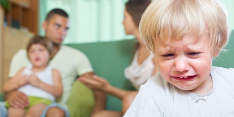Criança pequena chora em primeiro plano enquanto a família aparece desfocada ao fundo