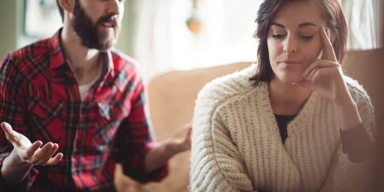 Mulher pensativa e cabisbaixa ao lado de homem que fala gesticulando no sofá, representando jogos mentais em uma conversa