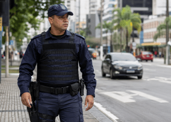 Guarda municipal de uniforme azul patrulha calçada de avenida movimentada, prédios ao fundo.