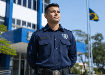 Guarda civil municipal de uniforme azul marinho com brasão, cinto com fivela, em frente a prédio azul e bandeira do Brasil.