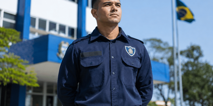 Guarda civil municipal de uniforme azul marinho com brasão, cinto com fivela, em frente a prédio azul e bandeira do Brasil.
