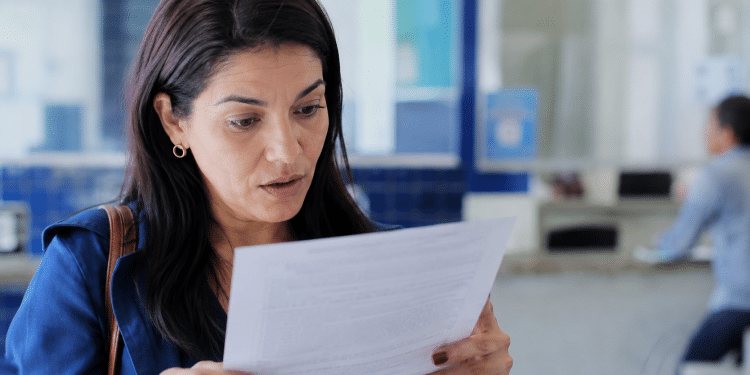 Mulher de cabelos escuros lendo papel com expressão surpresa em espaço interno.