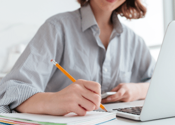 Jovem escrevendo com lápis em caderno ao lado de notebook, usando camisa listrada.