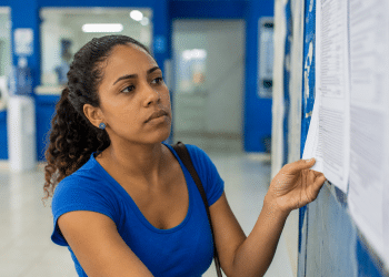 Mulher observa atentamente lista impressa afixada em mural azul em ambiente institucional.