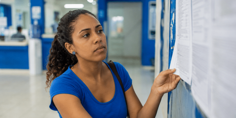 Mulher observa atentamente lista impressa afixada em mural azul em ambiente institucional.