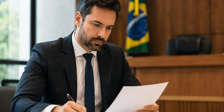 Homem de terno azul marinho e gravata assina documento em mesa de escritório, com bandeira do Brasil ao fundo.