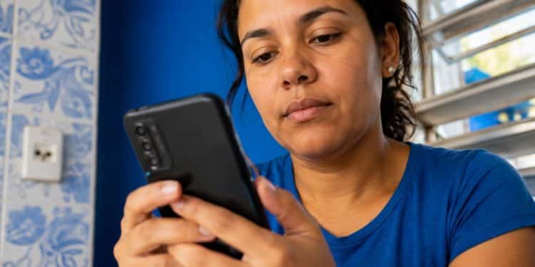 Mulher de camiseta azul consulta atenta a tela do celular para verificar a restrição de um aparelho pelo IMEI.