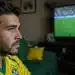 Jovem brasileiro assiste a jogo da Copa do Mundo na TV com expressão de cansaço visual.