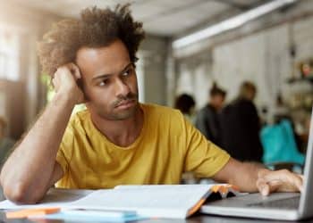 Jovem pensativo apoia a cabeça na mão diante de livro e notebook, refletindo sobre o futuro profissional
