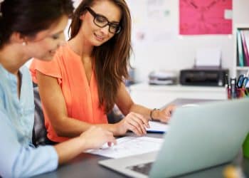Duas mulheres analisam documento e notebook juntas em uma secretaria escolar.