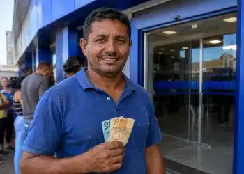 Trabalhador segura cédulas de real em frente a agência bancária, representando as datas de pagamento do PIS/Pasep e do INSS.