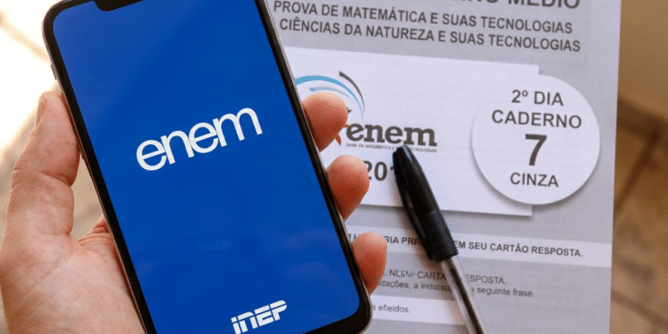 Mão segura celular com tela do ENEM ao lado de prova impressa e caneta preta.
