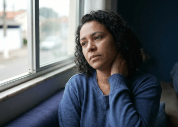 Mulher de expressão preocupada observa a rua pela janela enquanto apoia a mão na nuca, sinal de tensão