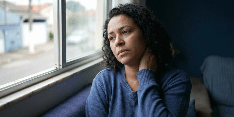 Mulher de expressão preocupada observa a rua pela janela enquanto apoia a mão na nuca, sinal de tensão