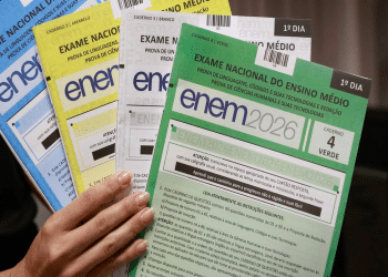 Mão segura quatro cadernos do ENEM 2026 em verde, branco, amarelo e azul com instruções visíveis, simbolizando os motivos que podem levar à eliminação no Enem.