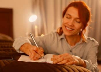 Mulher ruiva sorri ao escrever em diário na cama sob luz aconchegante de abajur à noite.