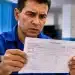 Trabalhador brasileiro observa contracheque impresso com expressão de dúvida, representando a incerteza sobre a forma correta de escrita da palavra.