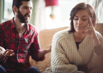 Mulher de suéter bege ignora homem de camisa xadrez vermelha que gesticula durante discussão no sofá, simbolizando frases que pessoas arrogantes costumam dizer com frequência.
