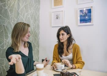 Duas mulheres sentadas à mesa de café, uma gesticulando enquanto a outra olha séria, parede decorada ao fundo.