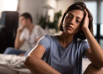 Mulher pensativa e abatida apoia a cabeça na mão enquanto homem aparece distante ao fundo no quarto