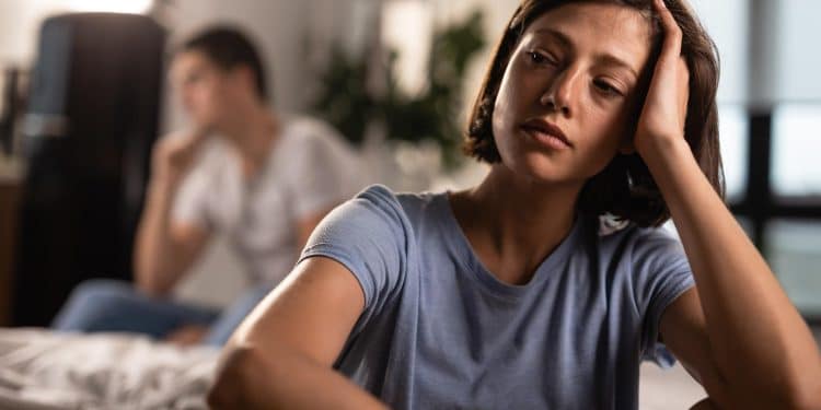Mulher pensativa e abatida apoia a cabeça na mão enquanto homem aparece distante ao fundo no quarto