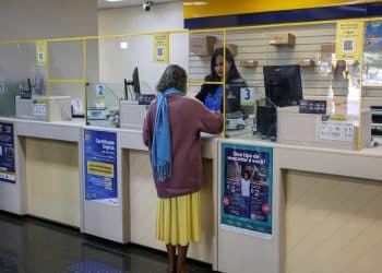 Mulher idosa de cabelo grisalho e cacheado, casaco roxo e cachecol azul, sendo atendida por funcionária em agência dos Correios, simbolizando o funcionamento dos bancos nos jogos do Brasil.