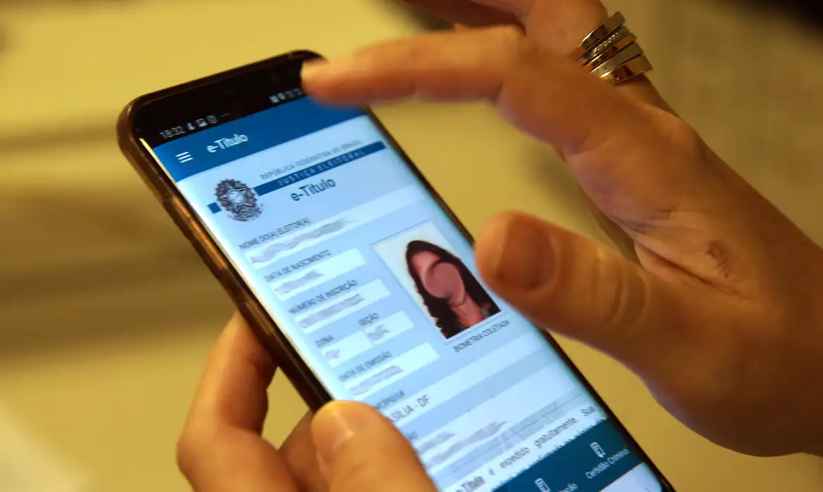 Pessoa segurando o celular com o e-Título aberto, mostrando foto, número de inscrição, zona e seção eleitoral
