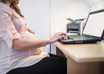 Mulher grávida sentada ao computador com uma mão na barriga e outra digitando, simbolizando o Projeto de Lei que propõe participação remota e segunda chamada para gestantes em concursos públicos.