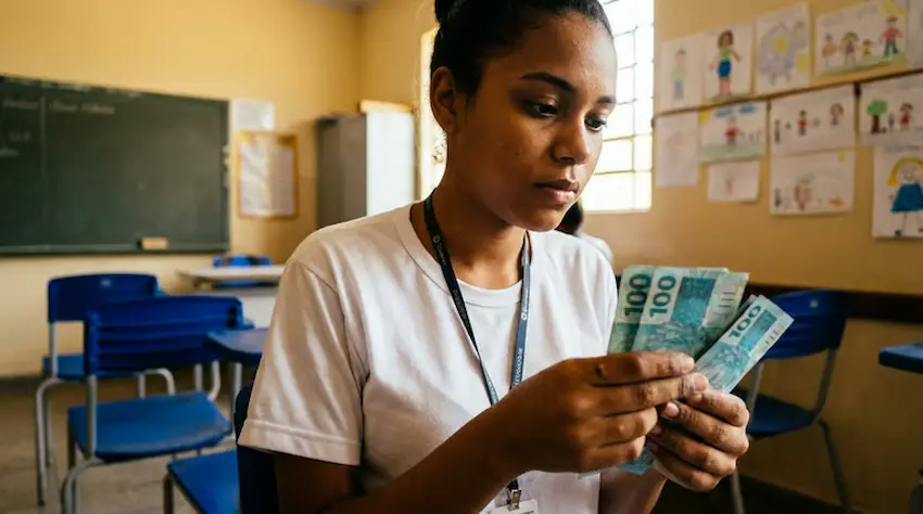 Estudante em sala de aula conferindo notas de R$100, relacionado ao pagamento do programa Pé-de-Meia de junho e julho.