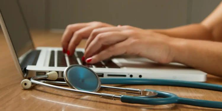 Mãos digitam em um notebook ao lado de um estetoscópio sobre a mesa, em imagem que ilustra a inscrição on-line do Enamed 2026