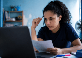 Jovem mulher assiste aula no notebook, segura folha e caneta, com expressão de foco e prateleira ao fundo.
