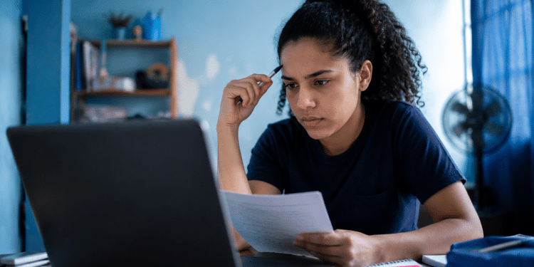 Jovem mulher assiste aula no notebook, segura folha e caneta, com expressão de foco e prateleira ao fundo.