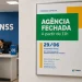 Interior de agência do INSS com atendente em guichê e placa informando fechamento antecipado do atendimento e alteração no horário de funcionamento do serviço 135.