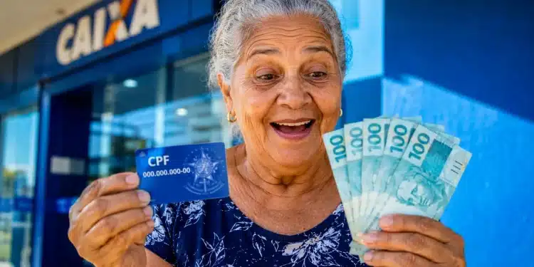 Idosa brasileira sorrindo segura cartão de CPF e notas de real em frente a uma agência bancária, representando os pagamentos de junho.
