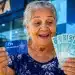 Idosa brasileira sorrindo segura cartão de CPF e notas de real em frente a uma agência bancária, representando os pagamentos de junho.