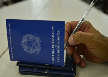 Mão masculina assinando Carteira de Trabalho azul com caneta em superfície branca, simbolizando o pagamento do lote extra do PIS/Pasep.