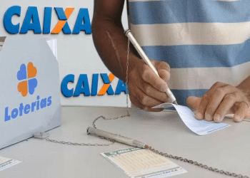 Pessoa de pele parda preenche bilhete da Mega-Sena ao lado de urna da Loterias Caixa, usando caneta presa por corrente, simbolizando a mudança de horário das loterias CAIXA.